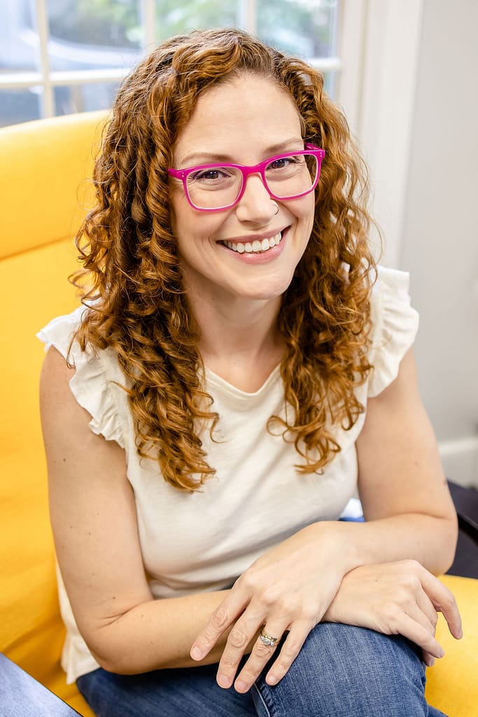 Rachel O'Brien, IBCLC sits in a yellow chair, with arms crossed, smiling at the camera